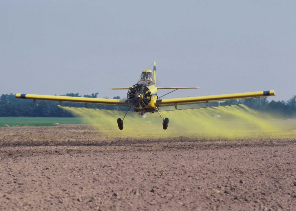 Avião agrícola pulverizando nuvem de agrotóxicos amarelos sobre o solo de uma lavoura.