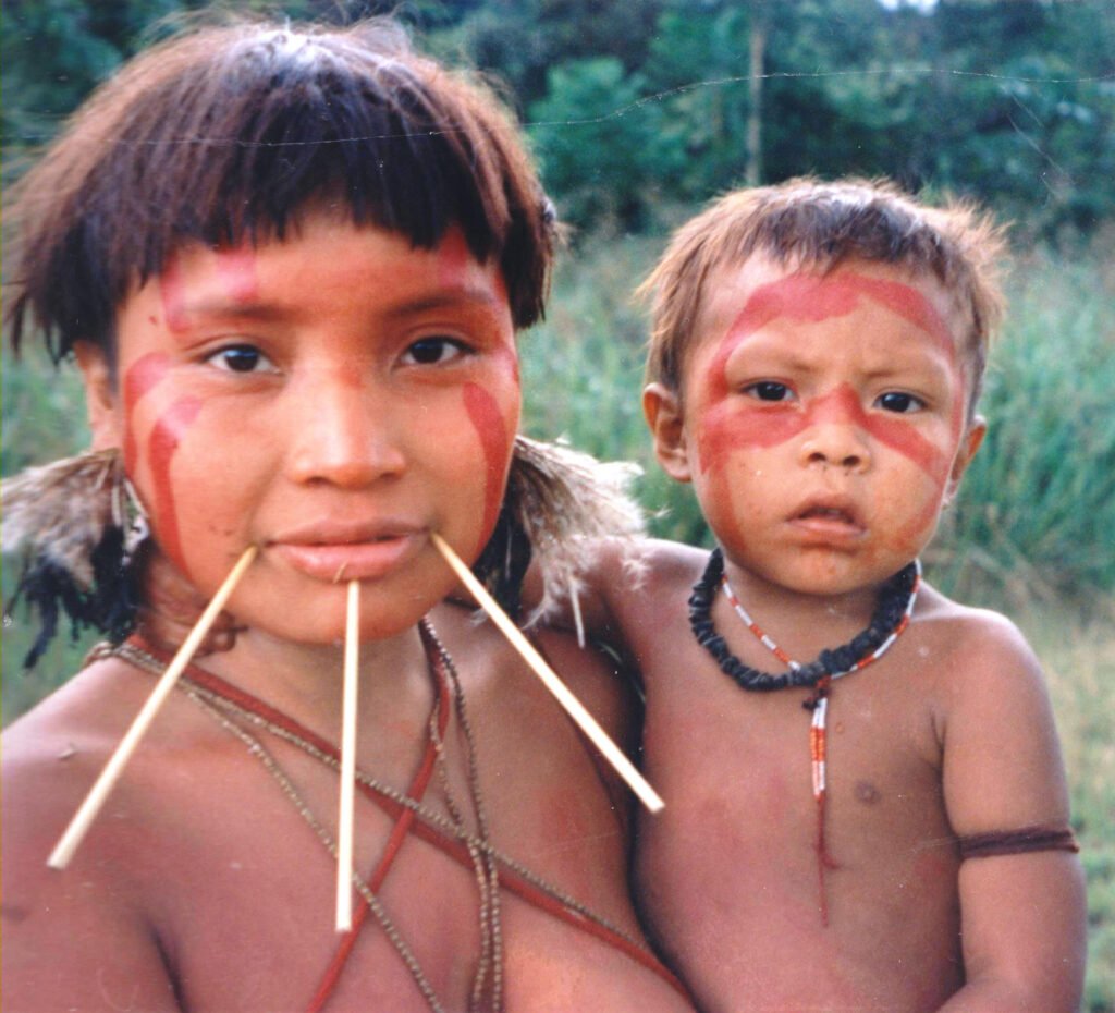 Mulher Yanomami com pintura vermelha e adornos tradicionais segura uma criança nos braços em meio à floresta amazônica.